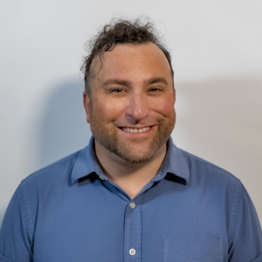 A person smiling at the camera in a professional headshot, wearing a light blue collared shirt, they stands against a plain light-colored background.