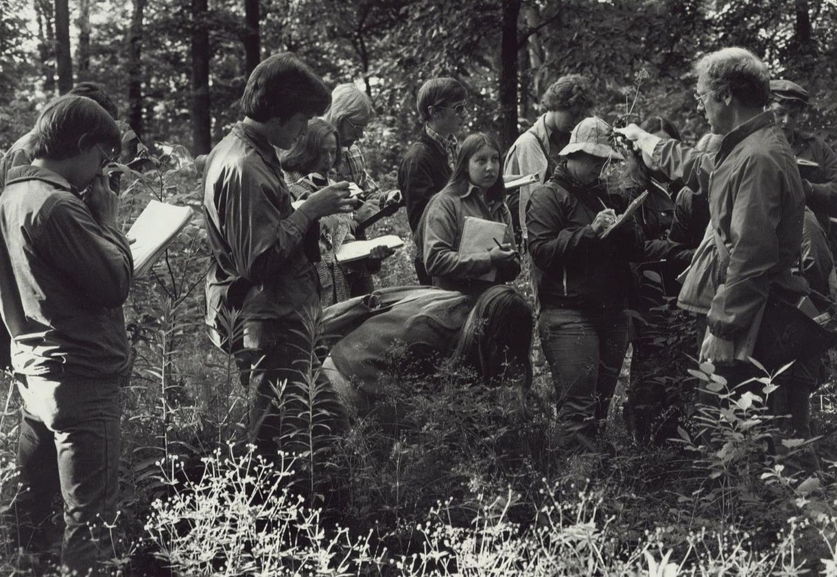 A black and white photo with a group of students, who are holding notebooks, listening to a professor. The students are outside in a forest and learning about plant life in southeastern Michigan