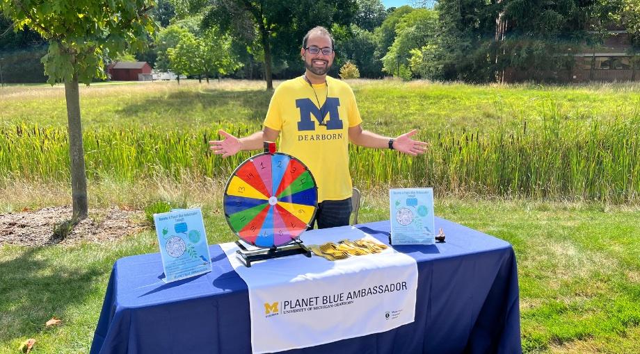 Student standing behind a Planet Blue Ambassador table wearing a UM-Dearborn maize shirt.