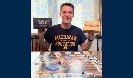 A photo of student Ezra Houghtby, sitting at a table in front of a Monopoly board while holding up boxes of two of his favorite board games.