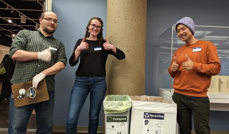 Three people give a thumbs up standing in front of compost and recycling bins