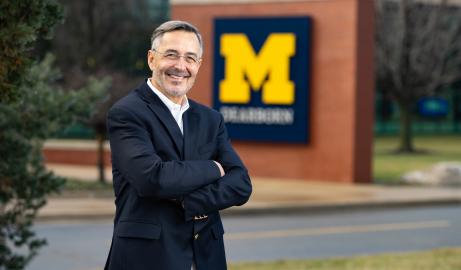 UM-Dearborn Chancellor Domenico Grasso, with his arms crossed and dressed in a blue sports coat, standing outside in front of a "Block M" UM-Dearborn logo