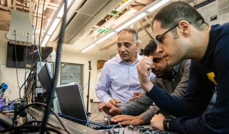 Professor Hafiz Malik, Associate Professor Alireza Mohammadi and a graduate student gather around stare at a laptop surrounded by a bunch of electronic components on a black work bench.