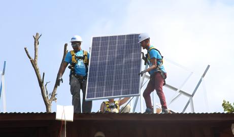 Two young men wearing hard hats and fall protection harnesses get ready to place a solar panel on the roof of a building