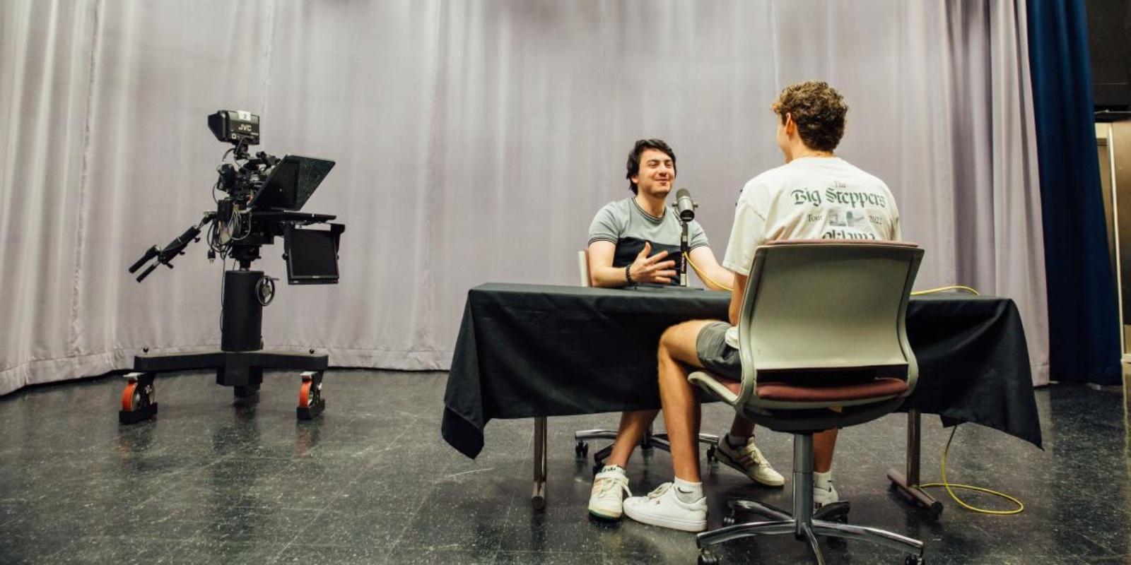 Two students record an interview inside a spacious recording studio with long gray drapes and a professional TV camera sitting in the background