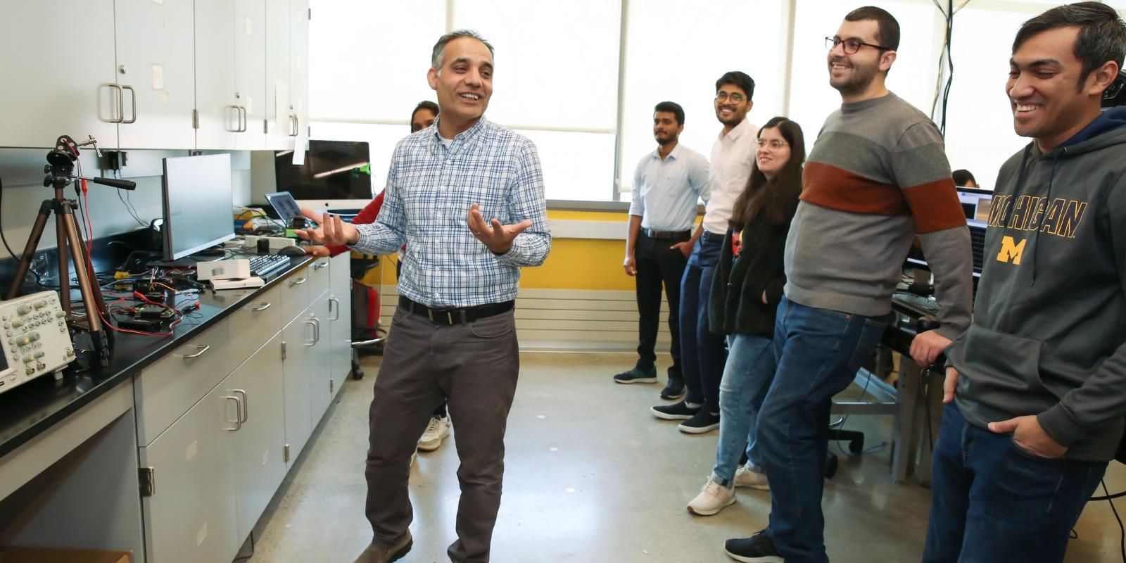Professor Hafiz Malik explains a concept to half a dozen graduate students, who look on smiling, in his lab at UM-Dearborn. 