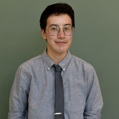 A light-skinned person with dark hair and glasses stands in front of a green background with their hands in their pockets. They are smiling slightly.