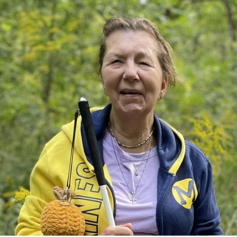 Donna posont smiles into the camera as she holds her cane, complete with a knit pumpkin. She is sporting a UM jacket standing outside by the Fair Lane Lake. 