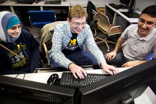 Three students gather around a gaming computer smiling and laughing