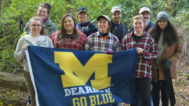 Group of students standing in forest area holding UM-Dearborn Go Blue flag.