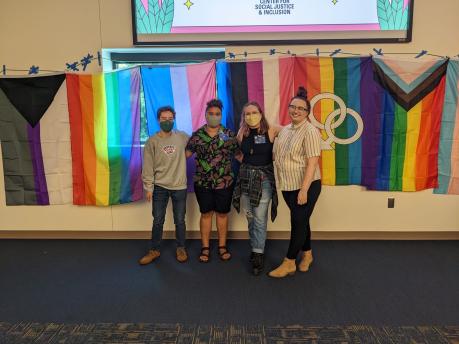 Four people stand in front of a white wall with various pride flags hung in a row. 