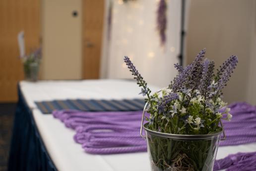 A white table is photographed, with a fake lavender plant in a see-through bucket in the foreground and several lavender graduation cords in the background (blurred).