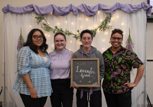 Four people standing in front of a white backdrop with lavender flowers and purple fabric hanging on top. The person in the middle-right is holding a sign that says "Lavender Grad"