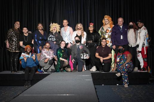 A large group of people are stood on a stage with black draping behind them. The group is comprised of mostly drag performers and some staff from the CSJI.