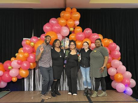 Five people are standing on a stage and smiling at the camera. There is a pink and orange balloon arch behind them.