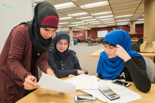 3 female students all wearing hijabs studying around a table. One is standing, two are sitting with papers and a calculator on the table.
