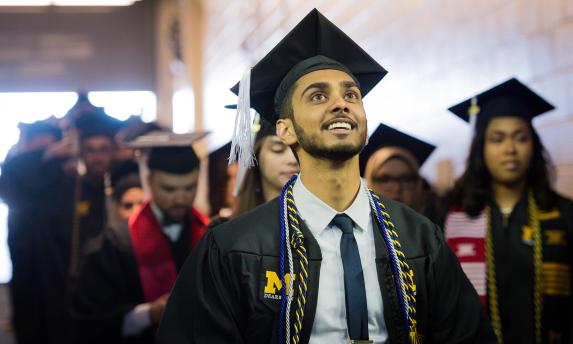 Student in academic gown smiling looking up at Commencement ceremony at Crisler Arena.
