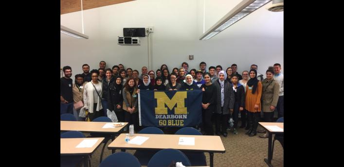 Group of people posed holding UM-Dearborn Go Blue flag inside.