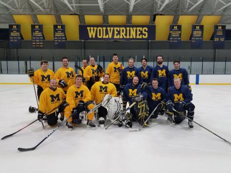 Alumni UM-Dearborn Hockey team pose for photo at mid-ice under Wolverines banner.