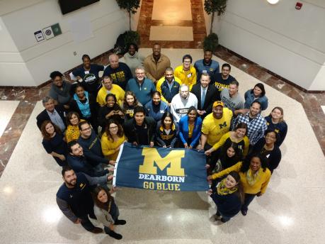 Alumni pose in atrium for photo from above holding a UM-Dearborn flag.