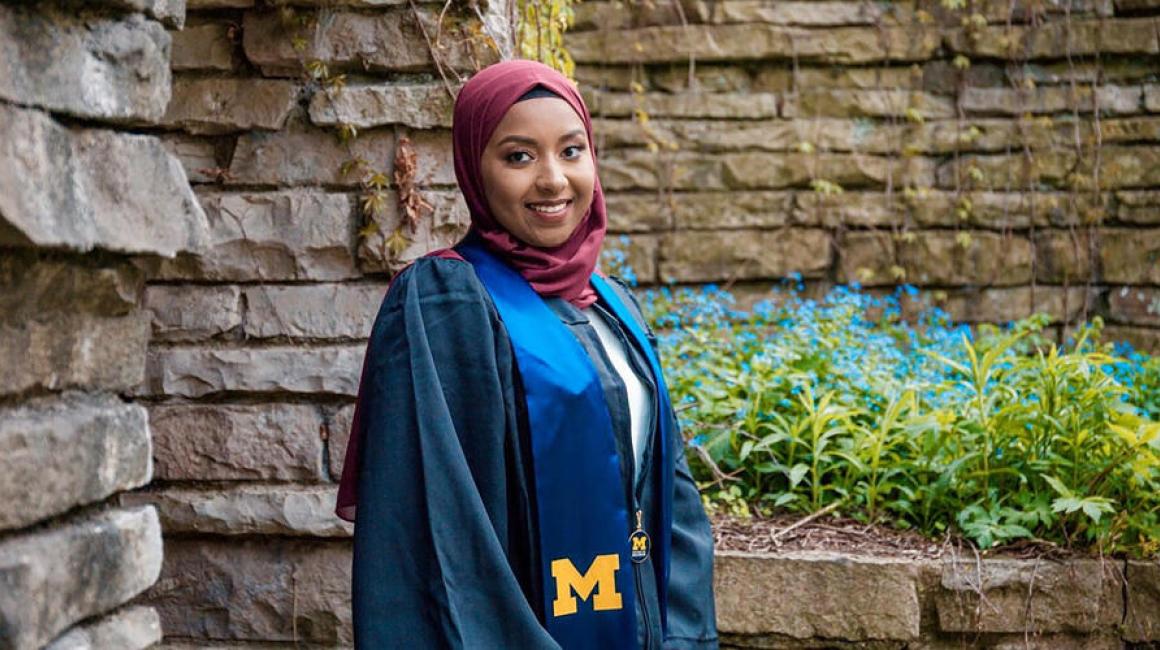 A portrait of recent UM-Dearborn grad Farzana Fariha in her commencement gown, standing against a stone wall covered in blue flowers.