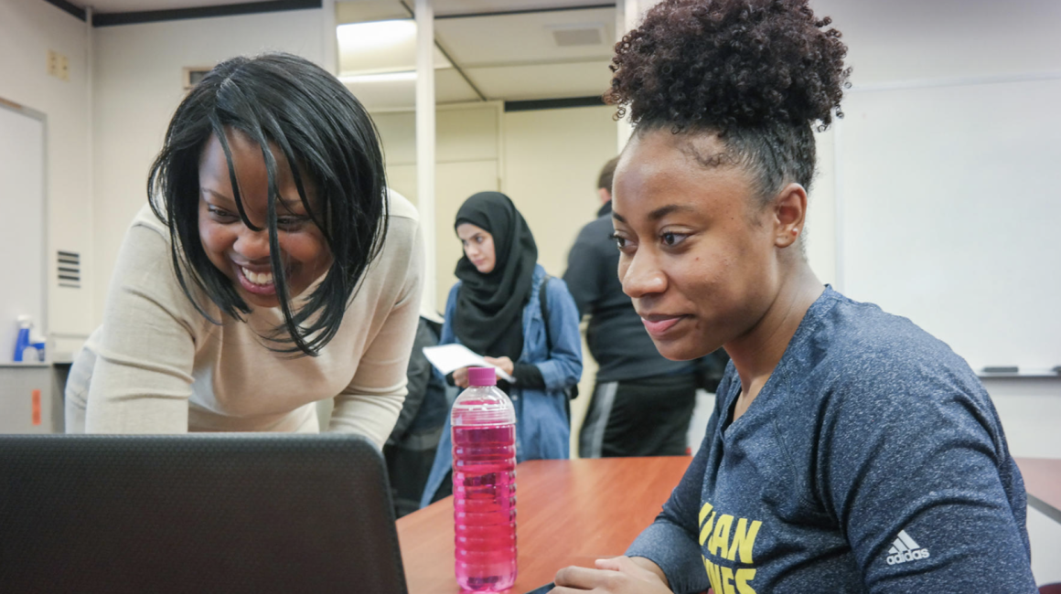 Bonnie McIntosh is a young Black woman with black, chin-length, straight hair. She is wearing a slim beige dress and is smiling and standing next to Sadé Lemons. Sadé is also a young Black woman with black, tightly coiled hair tied up. She is wearing a heather blue Wolverines shirt and is smiling, sitting looking at her laptop.