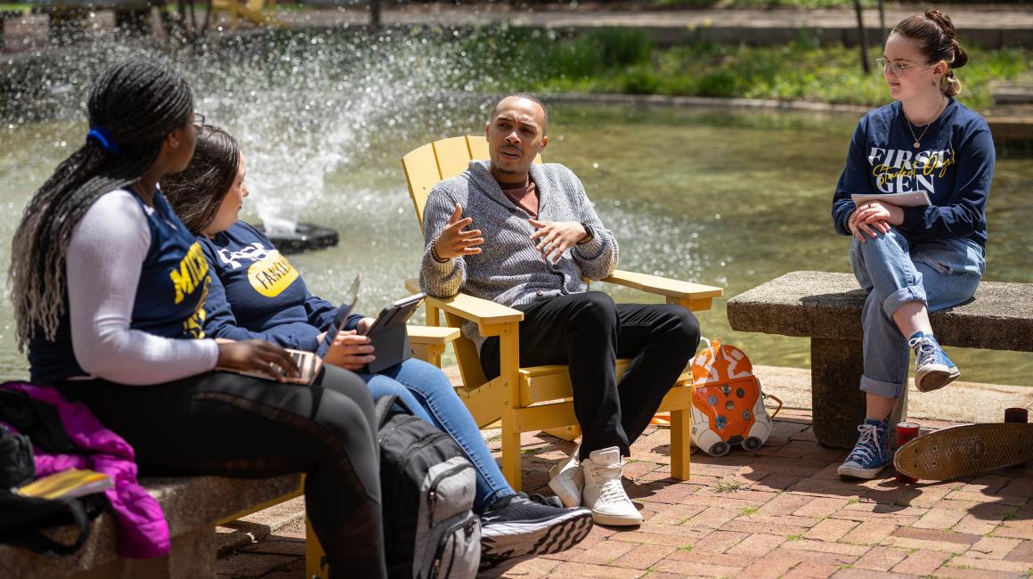 UM-Dearborn alum Maurice Taylor sits and talks with students by Chancellor's Pond with a fountain in the background