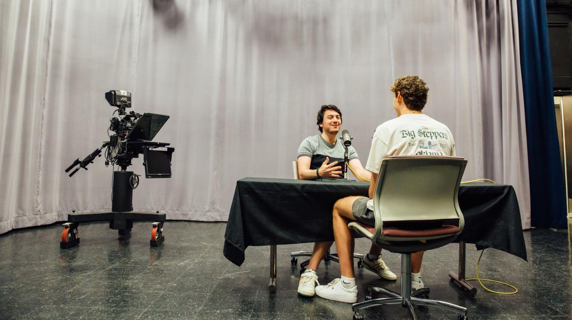 Two students record an interview inside a spacious recording studio with long gray drapes and a professional TV camera sitting in the background 
