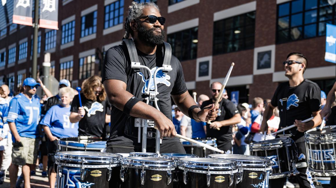 Wearing his six-drum marching kit, Alum DeAndre Hicks entertains Detroit Lions fans outside Ford Field on a sunny day.