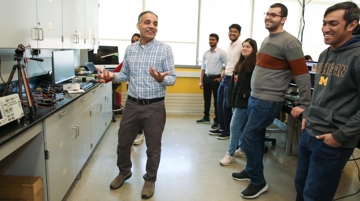 Professor Hafiz Malik explains a concept to half a dozen graduate students, who look on smiling, in his lab at UM-Dearborn lab