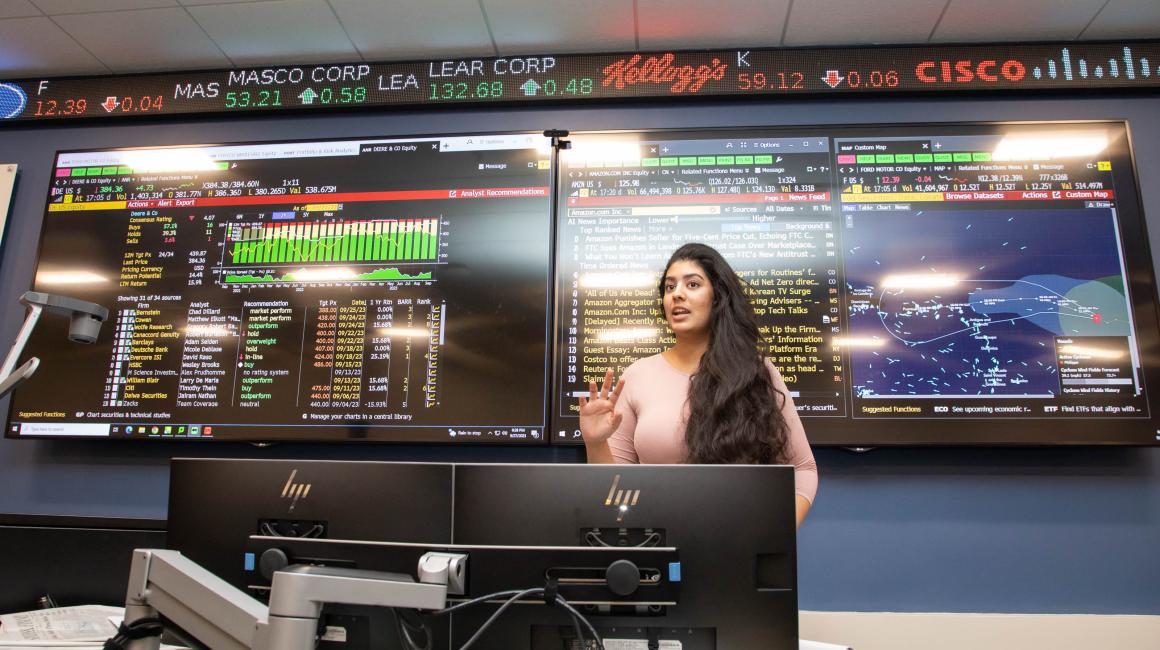 Set against a backdrop of stock tickers and business data displaying on two large LED screens, a graduate student speaks to a class in the Bloomberg Lab on the UM-Dearborn campus.
