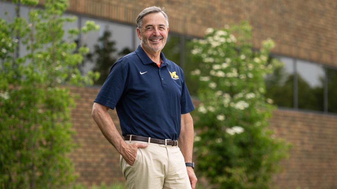 Chancellor Domenico Grass stands for a portrait in front of two small Dogwood trees in bloom on the UM-Dearborn campus