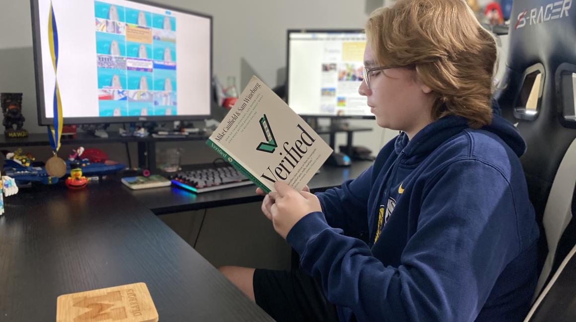 A student reading the book 'Verified' at a desk.