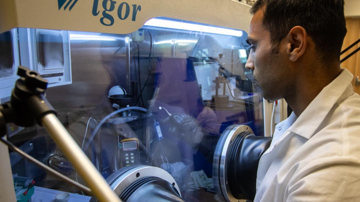 Wearing a white lab coat, graduate student Hossein Abbasi manipulates equipment inside a plastic chamber in the lab of battery researcher Assistant Professor Lei Chen.