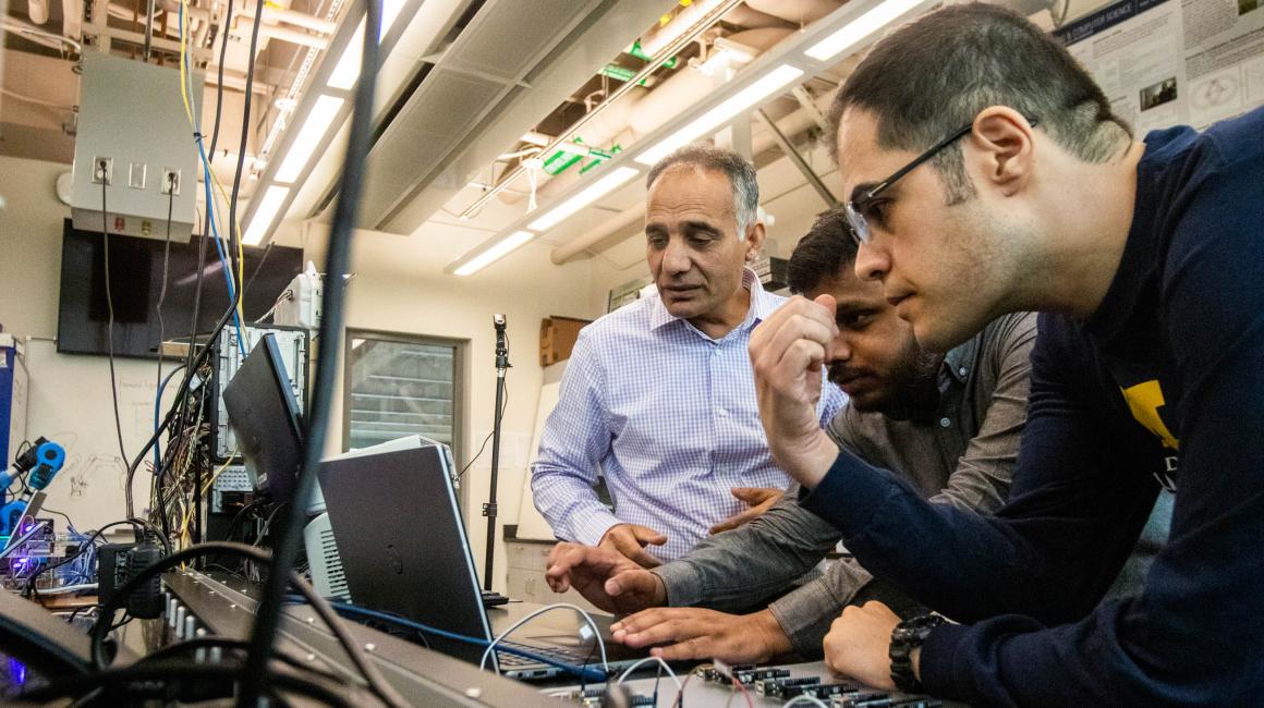 Professor Hafiz Malik, Associate Professor Alireza Mohammadi and a graduate student gather around stare at a laptop surrounded by a bunch of electronic components on a black work bench.