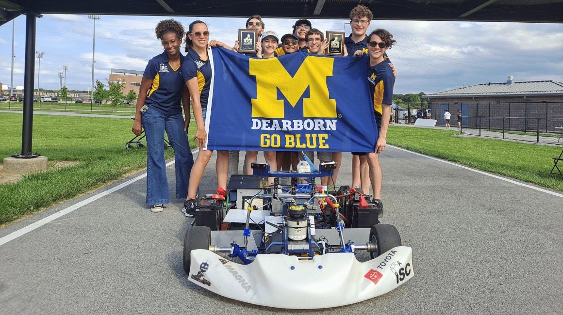 The Intelligent Systems Club stands on the race track holding a UM-Dearborn Flag