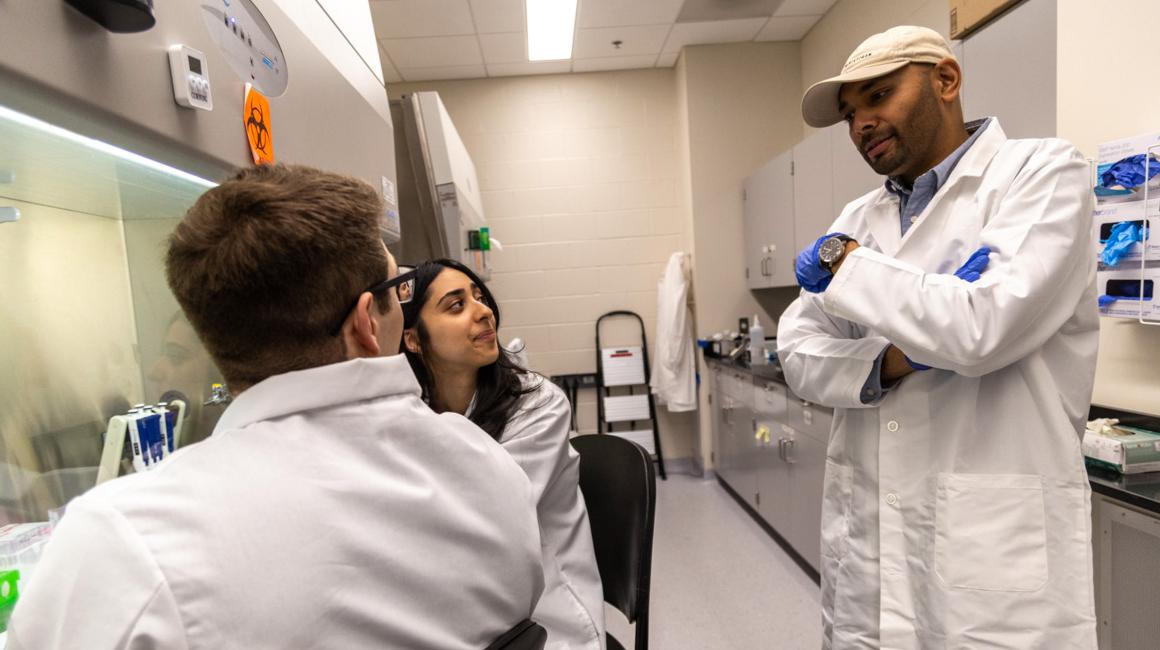 Wearing a white ball cap and white lab coat, Assistant Professor of Mechanical Engineering Aditya Raghunandan talks with two students in his bioengineering lab.