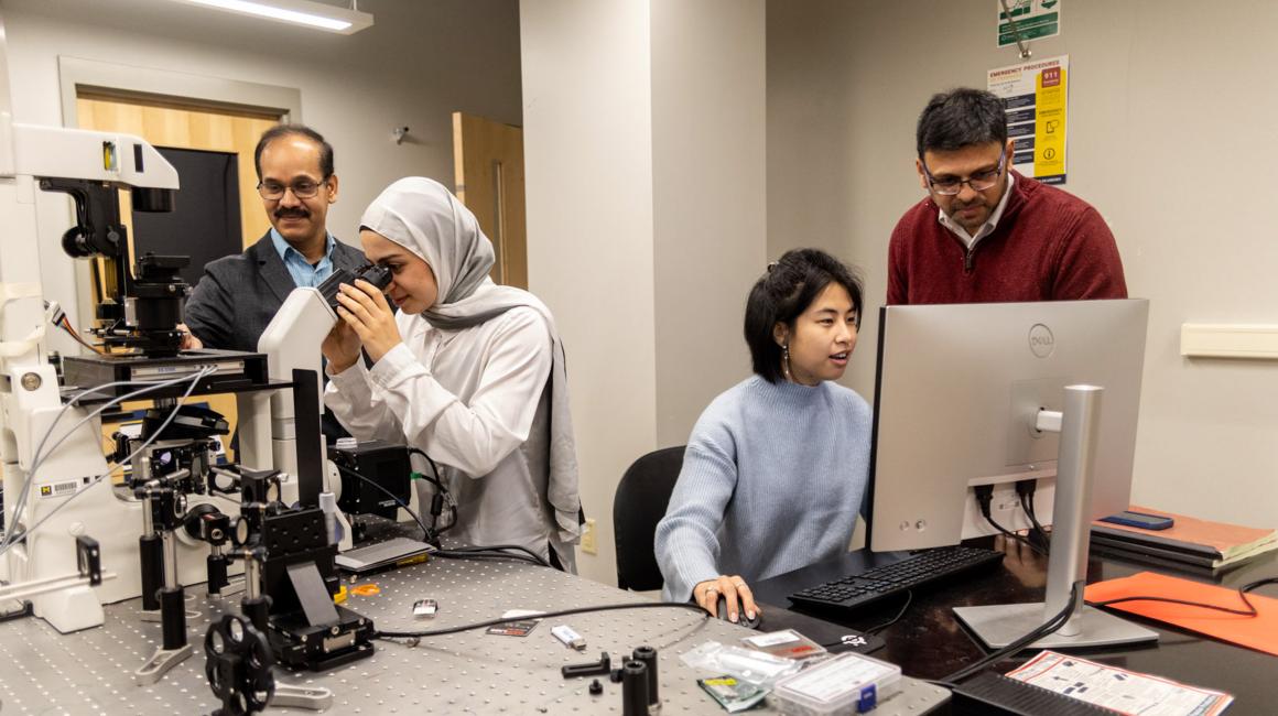 Flanked by microscopes and computers, Associate Professor of Biology Kalyan Kondapalli and Assistant Professor of Physics Suvranta Tripathy work with two female graduate students in a biology lab.