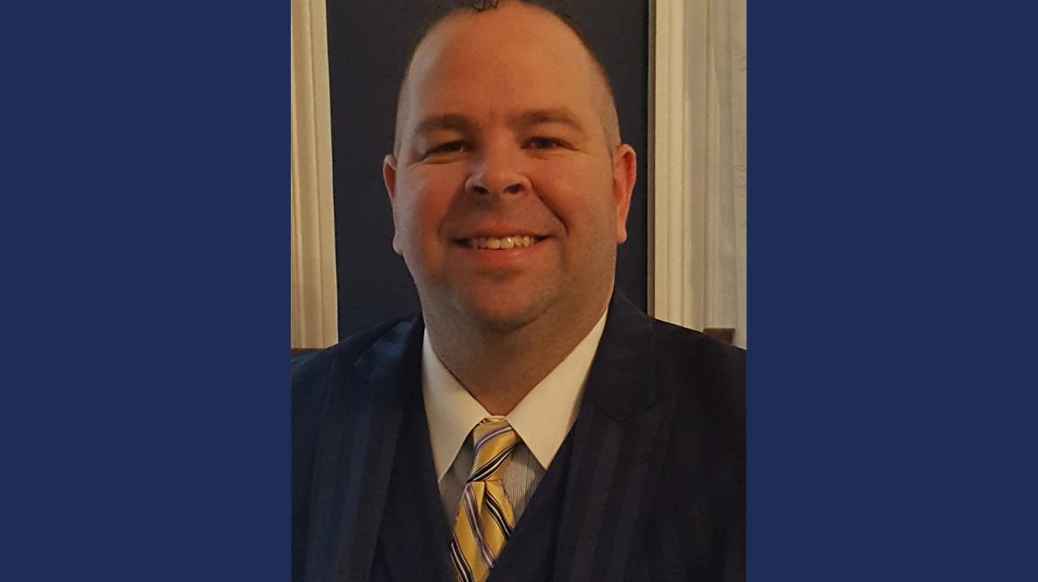 Captain Edward Hourigan is a middle-aged white man with thinning, brunet hair and brown eyes. He is wearing a navy suit jacket and vest over a white button up and maize & blue striped tie.
