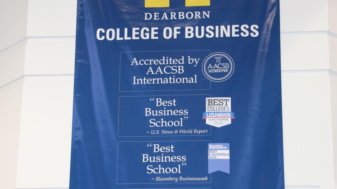 A long blue banner with the UM-Dearborn logo at the top and “College of Business” written underneath. The poster has seals for the AACSB, U.S. News Best Colleges, and Bloomberg Businessweek. It reads: “Accredited by AACSB International”, “Best Business School ~ U.S. News & World Report”, “Best Business School ~ Bloomberg Businessweek”