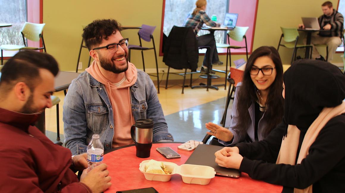 Mohamad Jaafar is a young, Arab man with brunet, curls and facial hair. He is laughing with a group of friends in the UC dining area. Mohamad is wearing a pair of glasses with a black top/wire bottoms and a salmon-pink hoodie with a jean jacket over.