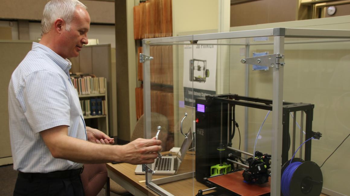 Patrick Armatis is an older white man with gray hair and no facial hair. He is wearing a short-sleeved blue and white gingham button down and black slacks. He stands in front of a 3D printer enclosed in an acrylic case.