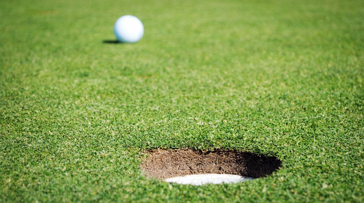 A stock image of a hole on a golf course. In the background, there is a white golf ball.