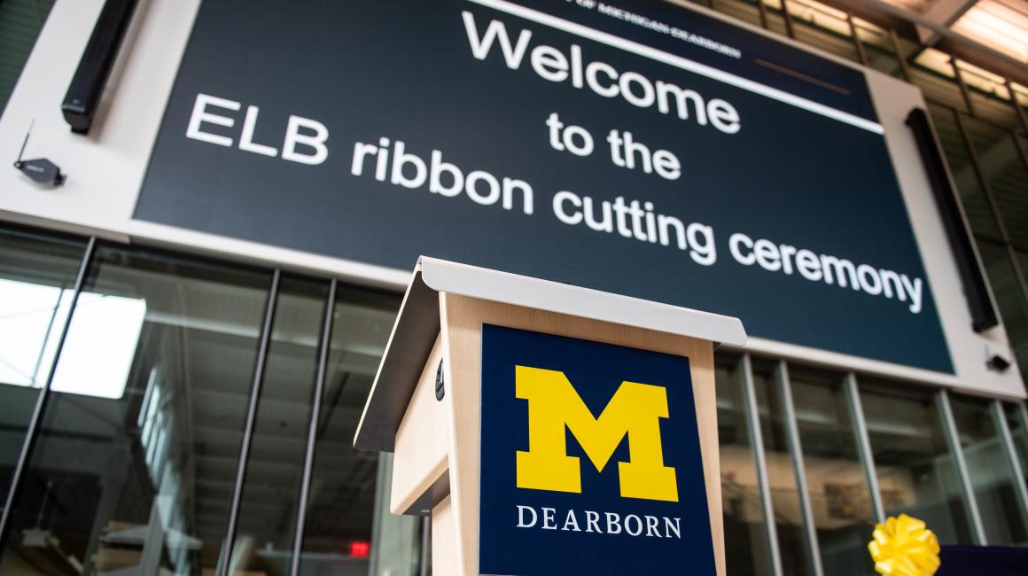 A shot of the screen inside of the Engineering Lab Building Atrium. In front of the screen is a podium with the UM-Dearborn logo. The screen reads, “Welcome to the ELB Ribbon Cutting Ceremony”
