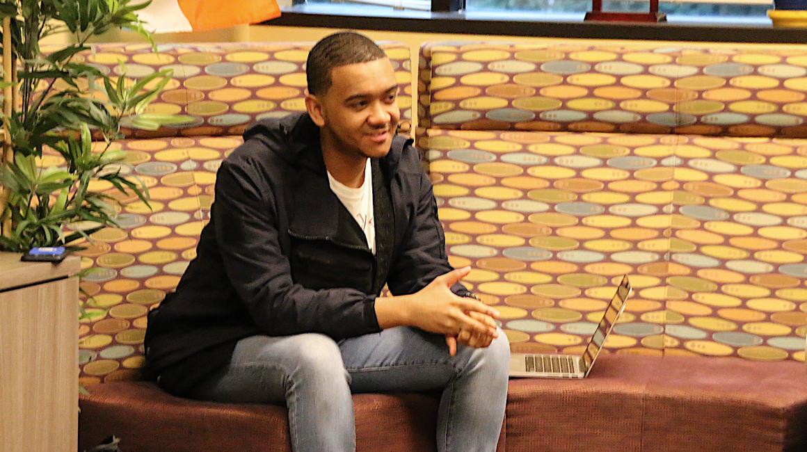 Adrian Maloy is a young Black man with brown short, faded waves and is wearing a white crew under a black jacket and light denim. He is sitting, facing away from the camera, on a red lounge sofa with his arms propped on his legs.