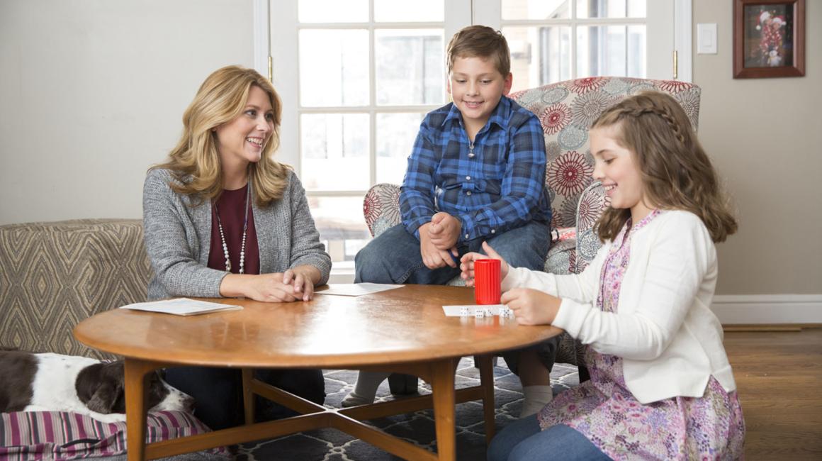 Andrea Ash sits at a table with her two children in a living space. Andrea is a middle aged white woman with shoulder-length blond hair. She is wearing a light gray cardigan with a reddish undershirt. Her son, to the immediate right of her is a white preteen boy with dark blond hair. He is wearing a blue plaid button down. Her daughter, to the far right is a white preteen girl with shoulder-length dark blonde hair. She is wearing a pink floral dress with a white cardigan.