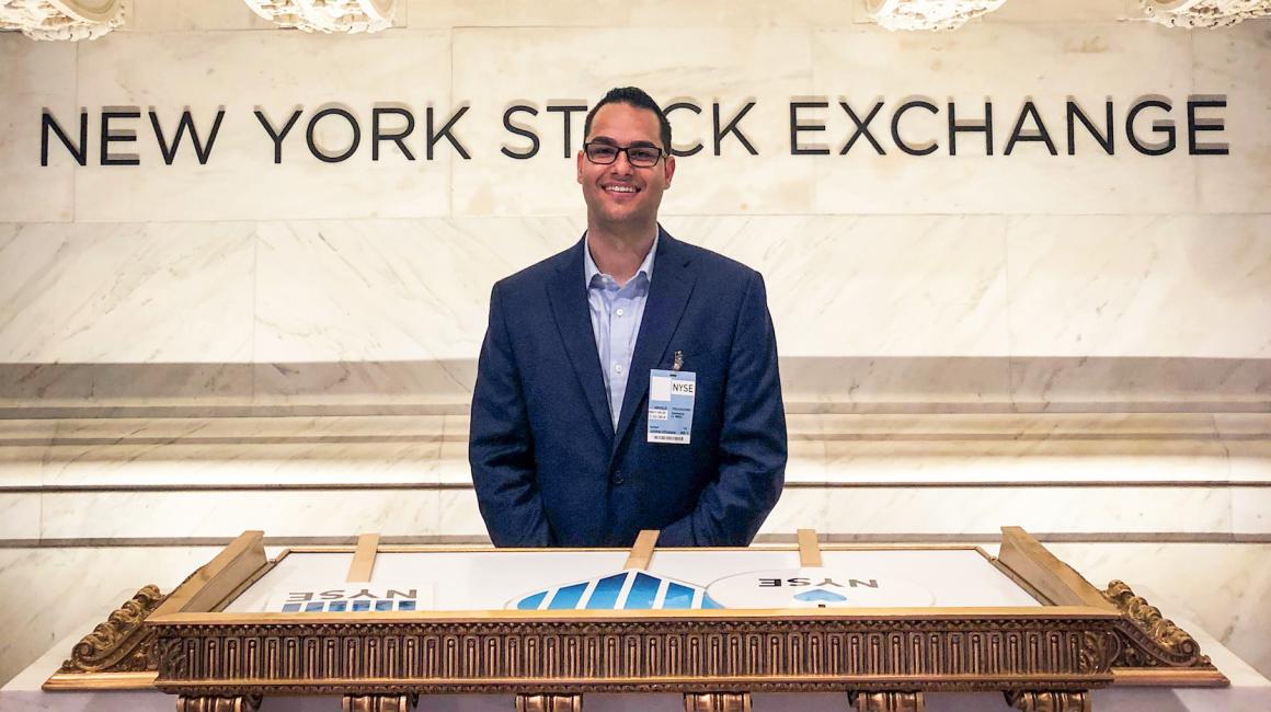 Angelo Policicchio is a young, Hispanic man with brown eyes and short, black hair. He is wearing a navy suit jacket over a powder-blue button down. Clipped onto his lapel pocket is a NYSE badge. Angelo stands in front of the entrance of the New York Stock Exchange.
