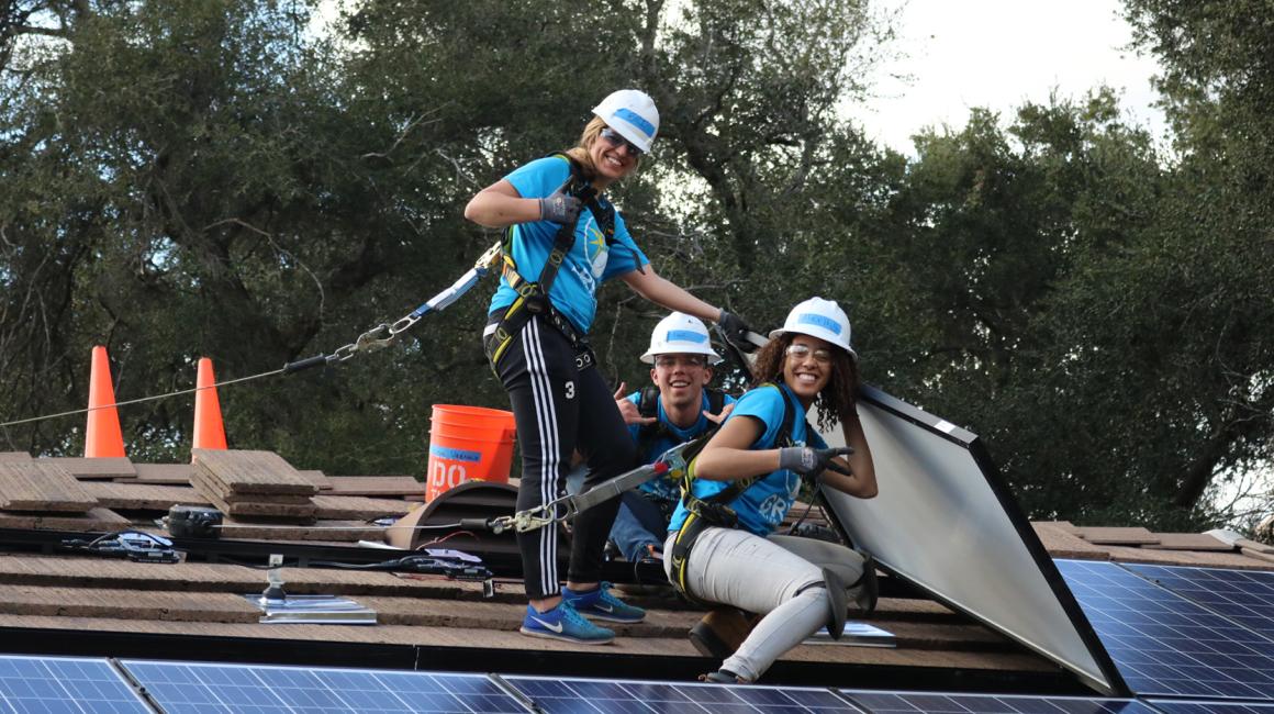 Two young, female students, and one young, male student sit on top of a roof, helping install solar panels.