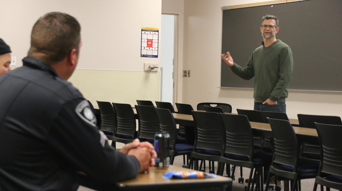 Professor Paul Draus stands in front of a class of officers. Paul is a middle-aged, white man with short gray hair and a goatee. He is wearing a pair of black, rectangular glasses, a green quarter-zip long sleeve and a pair of dark denim jeans.