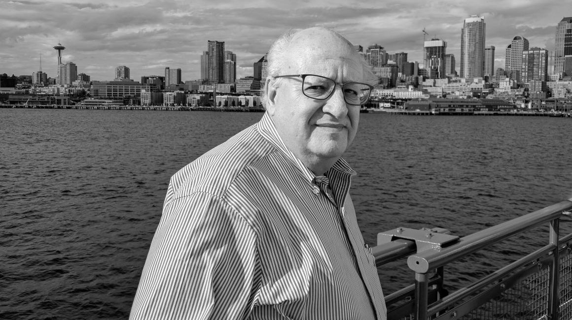 The late Professor Bill Grosky was an elderly white man with thinning gray hair and rectangular, black wire glasses. He was standing in front of the Detroit River, wearing a pinstripe button down. This photo is black-and-white.